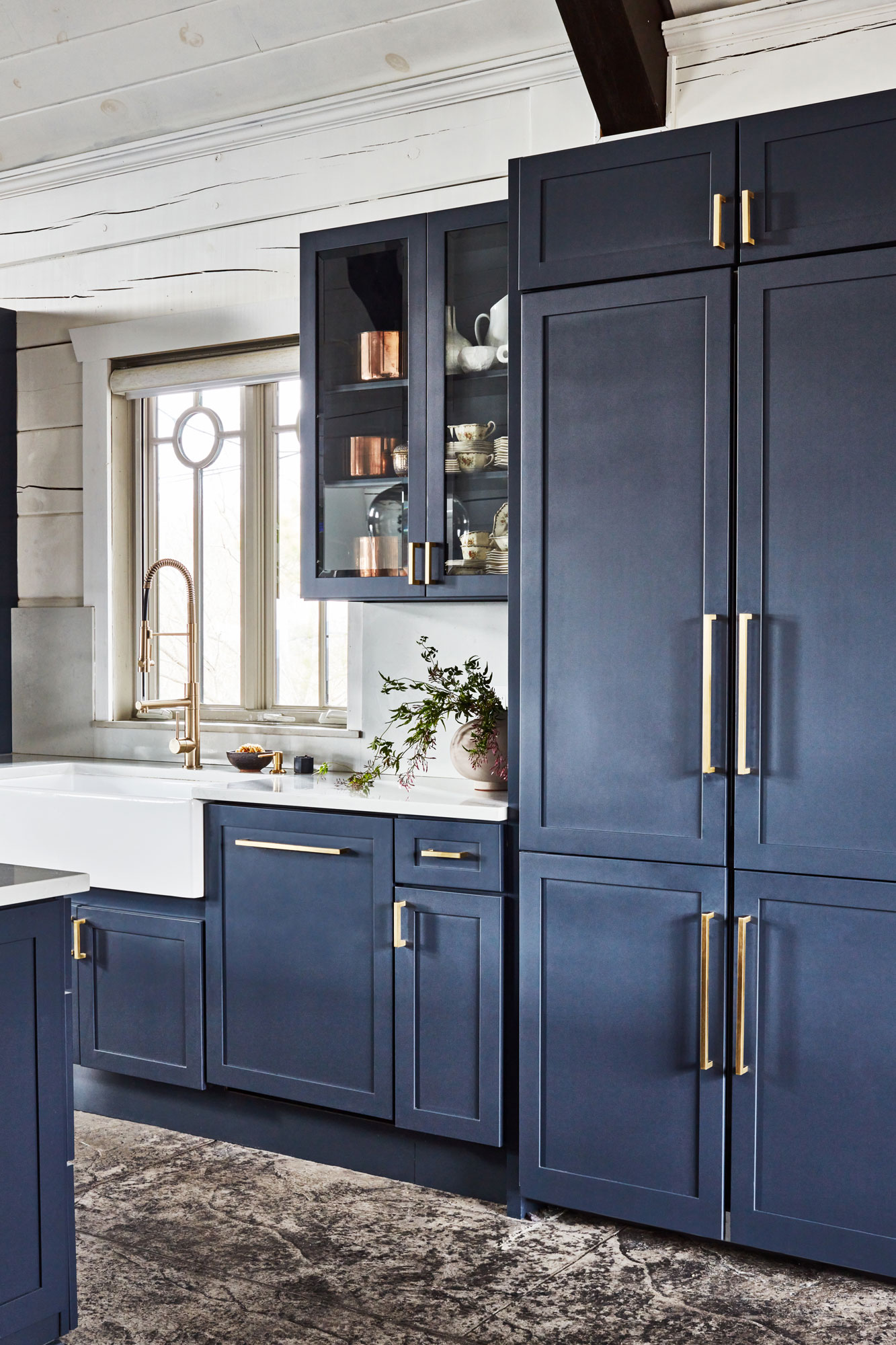 After the Remodeling: A modern kitchen with navy blue cabinets, brass handles, a farmhouse sink, and a brass faucet. A glass-front cabinet displays dishes; copper items sit on top. Light streams through a window above the sink, highlighting white walls and a rustic-style countertop.