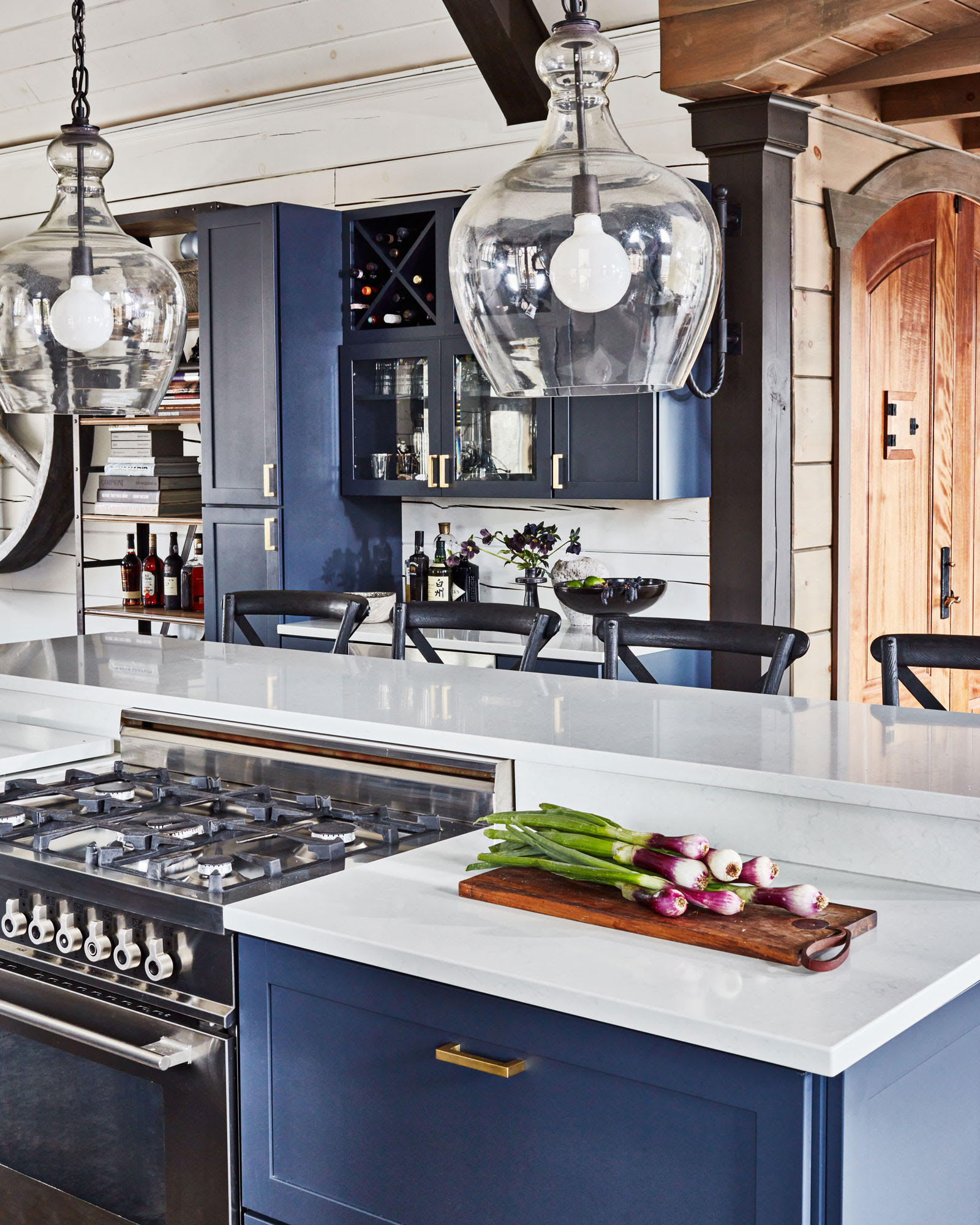After the Remodeling: Modern kitchen with blue cabinets, and a white countertop. A wooden cutting board with green onions and radishes sits atop the counter beside a stainless steel gas stove. Glass-front cabinets and wine storage are visible in the background.