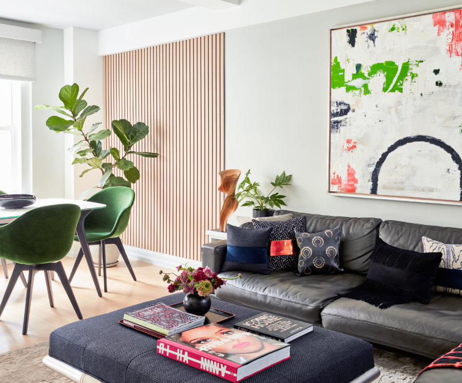A modern living room, crafted by Avery Johnson Design, features a gray sofa with patterned cushions, a navy ottoman topped with books and a flower vase, green dining chairs by a wooden table, lush potted plants, wood slat wall, and bold abstract art.