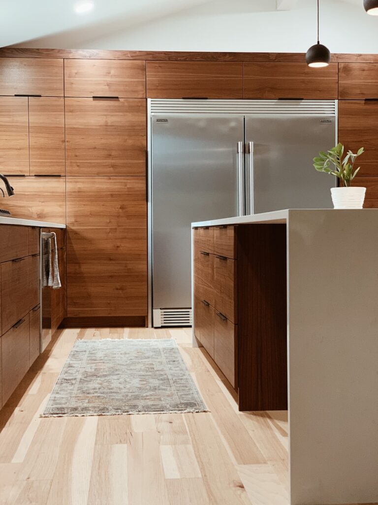 Modern kitchen with wood cabinetry & hardware design, light wood floors, and a large double-door stainless steel refrigerator. A small potted plant sits on the white island countertop, with a textured rug between the island and fridge.