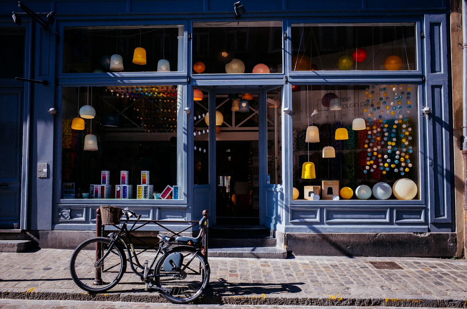 A bicycle is parked on a cobblestone street in front of a blue storefront showcasing thoughtful commercial interior design in Weehawken, with large windows displaying hanging lamps and colorful decor bathed in inviting sunlight.