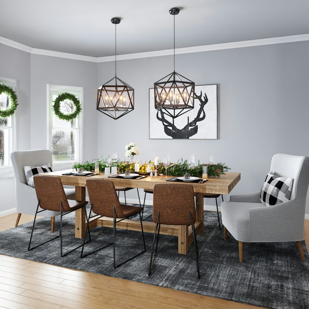 A modern dining room design features a wooden table set for a meal and six chairs—two as upholstered wingbacks. Two geometric pendant lights hang above. Wreaths adorn the windows, and a black deer silhouette artwork accents the gray wall.