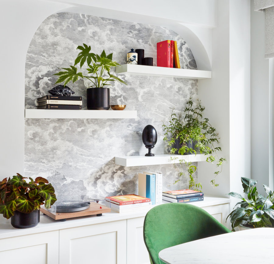 A modern nook, crafted by Avery Johnson Design, features two white floating shelves with books, black and white potted plants, decor accents, and a sculpture. Below, a built-in cabinet houses a turntable and books. A green velvet chair sits by a round table.