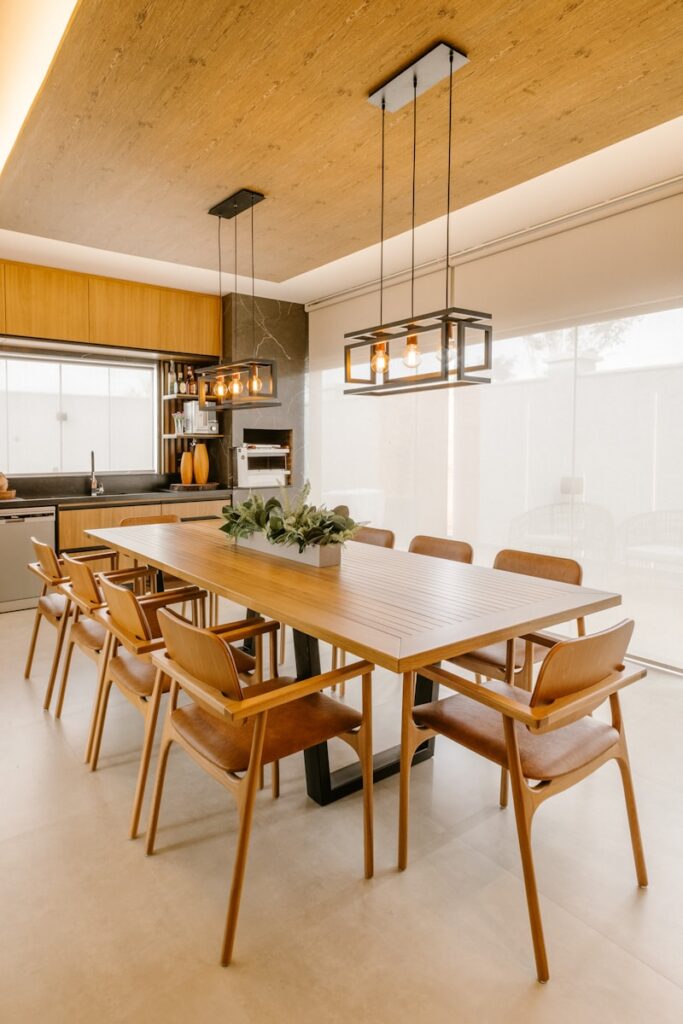 Modern dining room design featuring a long wooden table, ten matching chairs, minimalist decor, two rectangular pendant lights with exposed bulbs above, a sleek kitchen area with wooden cabinets, and large windows with sheer blinds letting in natural light.