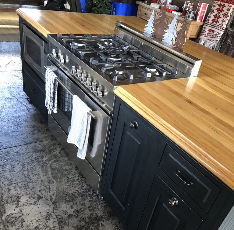 Before the remodeling: A kitchen island with a wooden countertop features a stainless steel gas stove and oven, dark cabinetry, drawers, and a towel hanging from the oven handle.